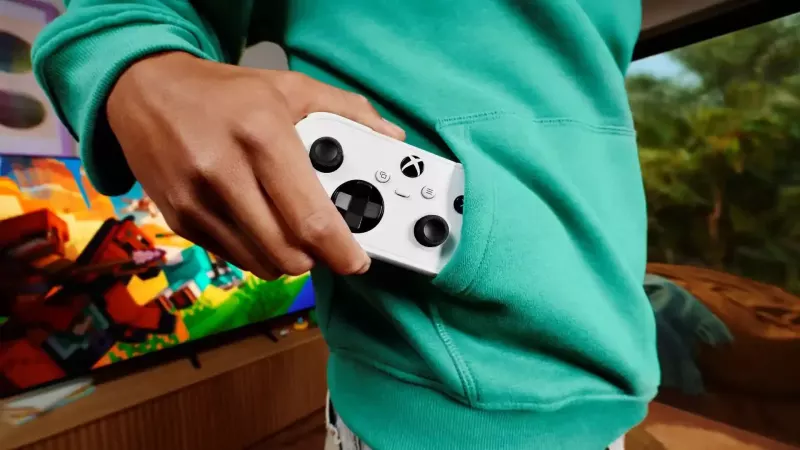 Osoba u tirkiznom duksu drži beli Xbox Nano kontroler delimično u prednjem džepu, dok se u pozadini vidi televizor sa igrom.