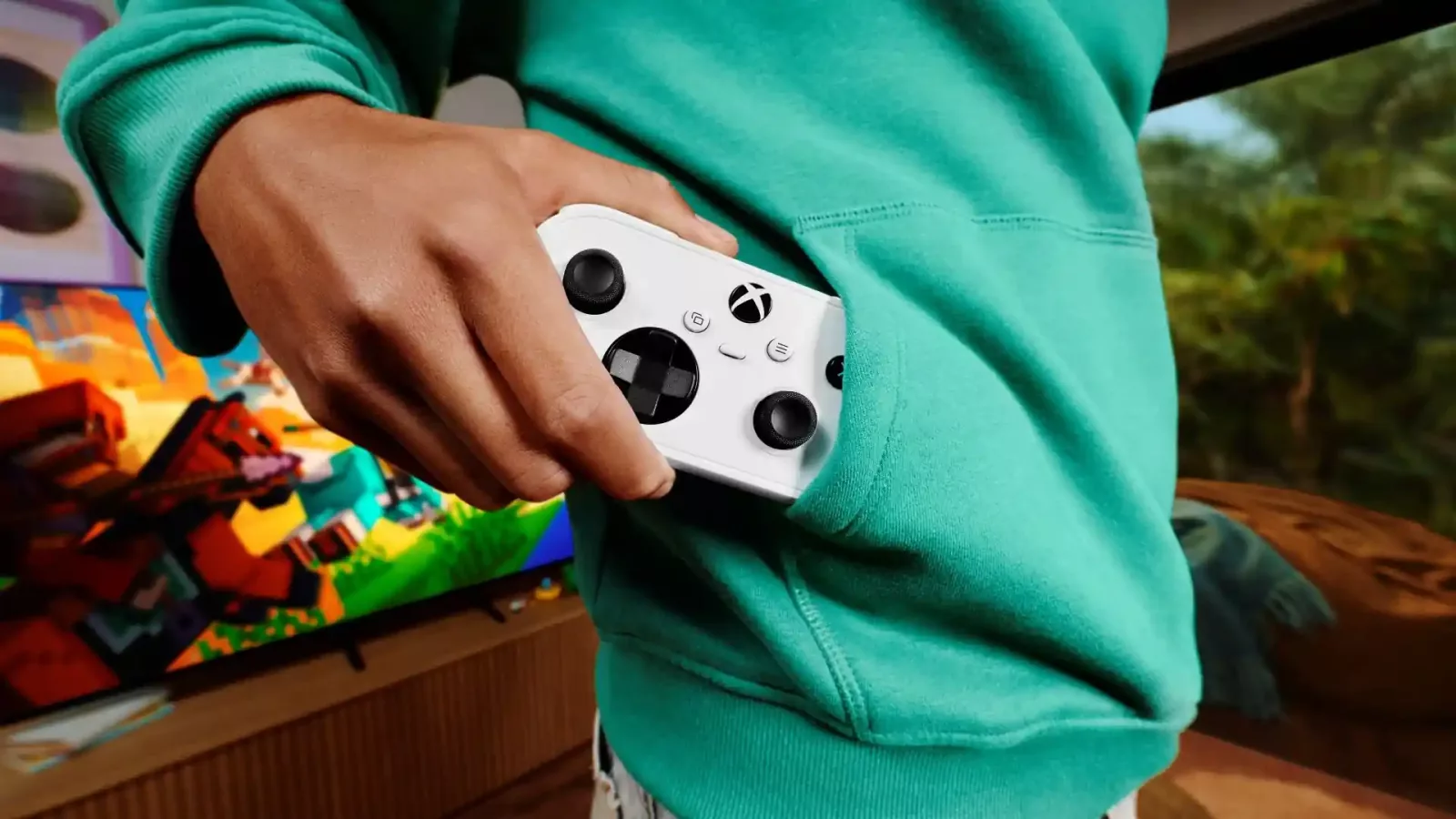 Osoba u tirkiznom duksu drži beli Xbox Nano kontroler delimično u prednjem džepu, dok se u pozadini vidi televizor sa igrom.